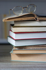 Stack of books on the table, reading glasses and e-reader on the table. Bookshelf in the background. Selective focus.