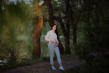 Fototapeta premium Sexy sensual brunette walks in a summer forest at sunset in a white blouse and jeans