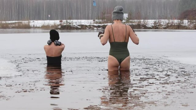 Two young, beautiful women take an ice bath in a frozen lake, surrounded by snow-covered nature. The icy water providing a refreshing and invigorating experience during winter swimming