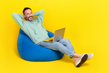 Full length photo of pretty dreamy man wear green sweatshirt hands arms behind head modern gadget beanbag isolated yellow color background