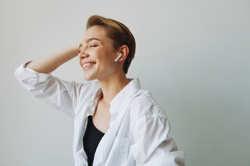 Young woman teenager listening to music with infertile headphones and dancing home, grinning with teeth with a short haircut in a white shirt on a white background. Girl natural poses with no filters