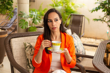 Uma jovem mulher tomando chá com expressão contemplativa, na varanda de sua casa.