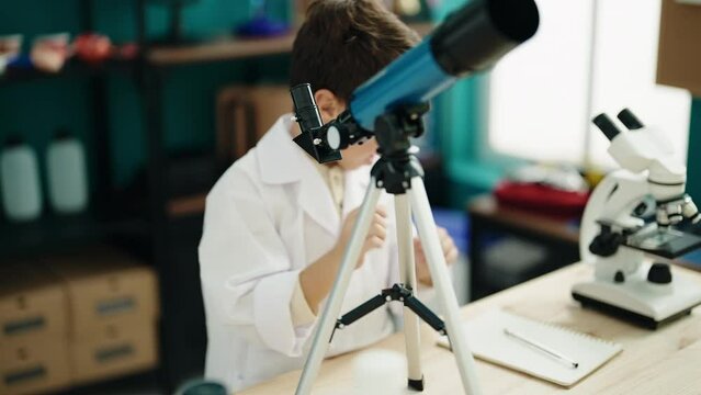 Adorable hispanic boy student using telescope writing on notebook at laboratory classroom