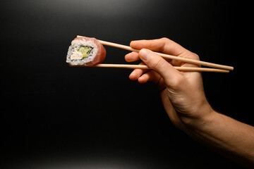 great side view of Philadelphia roll in hand holding with bamboo chopsticks on dark background.