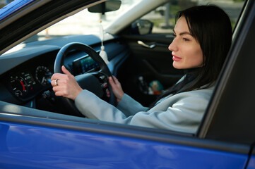 Fototapeta na wymiar Close-up side portrait of an attractive confident successful Caucasian businesswoman, experienced female driver with hands on steering wheel, driving her modern luxury car. People and Urban lifestyles