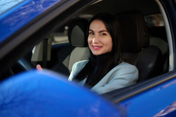 Attractive middle-aged dark-haired European successful woman, in formal wear, driving car and...