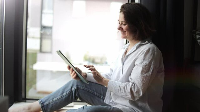 Business woman works in morning at home on tablet by window. Adult female freelancer, designer, programmer, financier