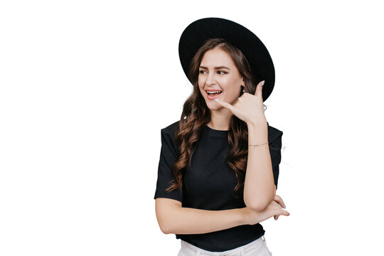 Young brunette hispanic girl in black hat with brim makes call gesture by hand looks aside at copy space over transparent background. Cheerful caucasian young woman with wavy hair. Promo, fashion.