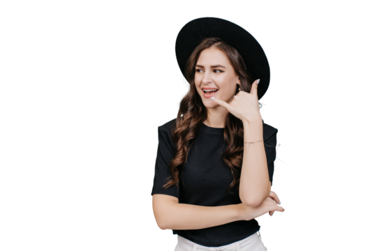 Young brunette hispanic girl in black hat with brim makes call gesture by hand looks aside at copy space over transparent background. Cheerful caucasian young woman with wavy hair. Promo, fashion.