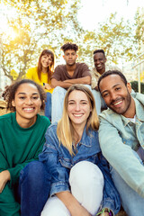Vertical shot of young student people smiling at camera sitting outdoors. Group photo of millennial friends laughing together resting in city street.