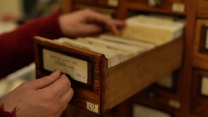 Woman worker looks for handwritten card going over big catalog to find book records in vintage wooden drawer at old library closeup