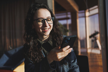 A cute woman works on a laptop in a cafe. Young concentrated brunette woman in glasses sitting at the table near the window drinking coffee. Freelance and remote work. Modern women's lifestyle