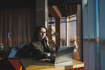 Young freelancer woman. Young woman working with laptop in coworking space and talking on phone, sitting in open space with cup of coffee and smiling while holding glasses.