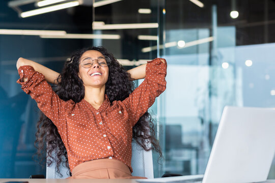 A Young Beautiful Latin American Woman Student Sits In The Office, Campus At A Table With A Laptop, Put Her Hands Behind Her Head, Closed Her Eyes, Rests From Studying, Smiles.