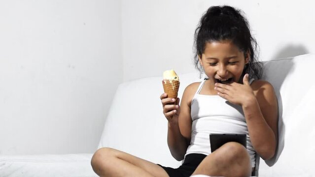 brunette latina girl, eating ice cream and looking at her tablet, amazed and impressed with what she is seeing, sitting on the sofa at home.