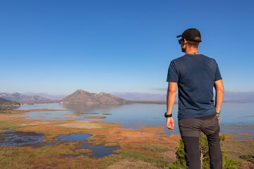 Rear view of man with panoramic view of Lake Skadar National Park in autumn seen from Virpazar, Bar, Montenegro, Balkans, Europe. Stunning travel destination in Dinaric Alps near Albanian border - obrazy, fototapety, plakaty