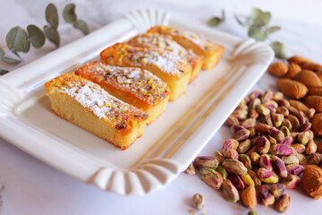 Financieros de almendra con pistacho y azúcar glass por encima en fuente de cerámica imitando bandeja de cartón blanco de pastelería, sobre mesa de mármol y junto a almendras con cáscara y pistachos