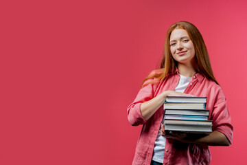 Clever student holds stack of university books from college library on pink background. Happy red-haired girl smiles, she is happy to graduate. Copy space.