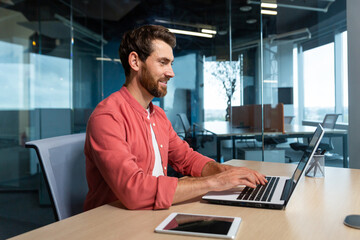 Successful businessman in red shirt happily working with laptop inside office, mature man with beard at workplace typing on keyboard smiling satisfied with work results and achievement.