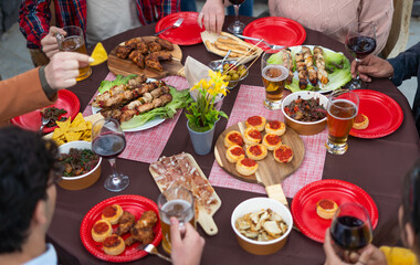 A beautifully set table featuring an array of delicious grilled meats, mini pizzas, crispy fries alcoholic beverages. Multiracial group of friends having summer party outdoor