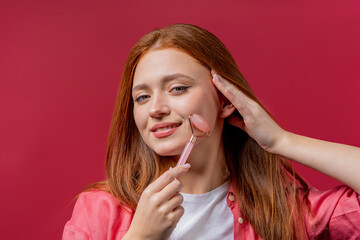 Ginger woman doing face massage with rose quartz stone roller on pink background. Facial self care,...