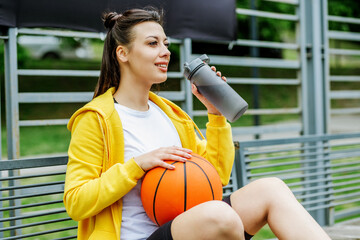 Portrait of young woman sitting on bench with basketball and drinking water during break