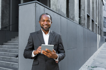 Portrait of a successful African American boss, a businessman in a suit smiling and looking at the camera, a man holding a tablet computer in his hands, a successful investor browsing the Internet.