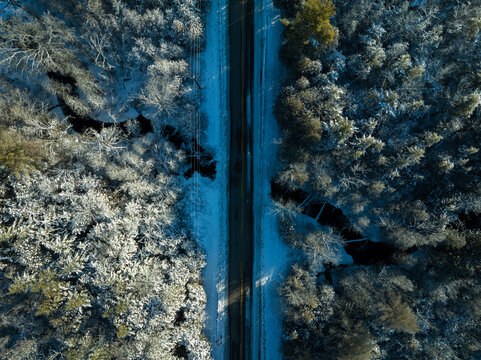 A high aerial photo centred above a two-lane, straight road passing over a creek in a rural area and running through a frozen, snow-covered forest in the winter.