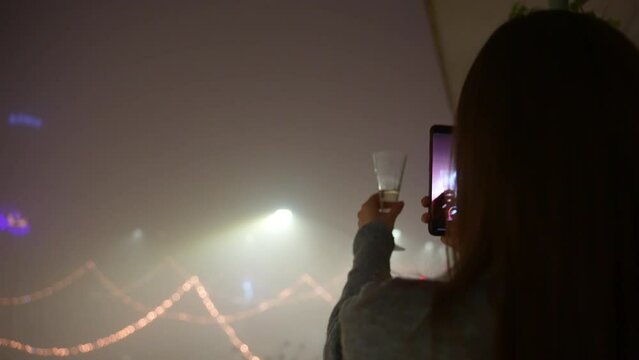 Woman taking photos of night foggy street and champagne glass on cellphone, beautifully decorated and illuminated night street at new years eve party or Christmas. Festive photography taken on