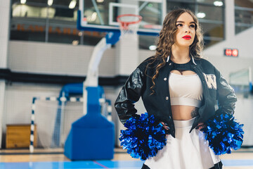 Horizontal shot of cheerleader wearing red lipstick in a jacket posing with blue shiny pom-poms. Basketball court blurred in the background. High quality photo