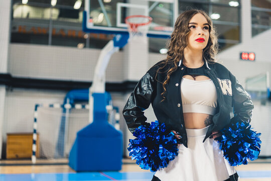 Horizontal shot of cheerleader wearing red lipstick in a jacket posing with blue shiny pom-poms. Basketball court blurred in the background. High quality photo