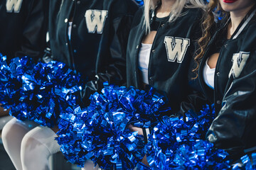 Close up shot of cheerleaders in black jumpers sitting in a row and holding blue, shiny pom-poms on...