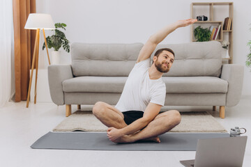 Active young cheerful man doing morning gymnastic at home, sitting on yoga mat and stretching, copy space