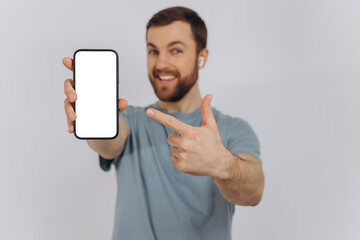 Cheerful Emotional Bearded Man Holding Blank Cell Phone In Hand Showing White Screen To Camera Pointing At You, Happy Millennial Guy Recommending New Application Or Mobile Website, Mockup Banner