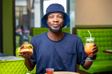 image of cheerful african guy holding sandwich and a glass of cocktail in a cafeteria- food concept