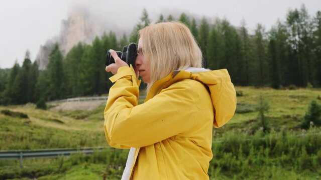 Blonde lady makes photos of meadows of Alps. Young woman tourist captures moments with camera and explores foggy hillside in mountains