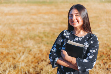 Fototapeta na wymiar Young smiling christian woman holding a bible under her arm in the field. Sola scriptura. Copy space