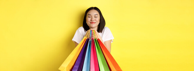 Obraz na płótnie Canvas Happy asian female shopper smiling and holding colorful shopping bags, standing against yellow background
