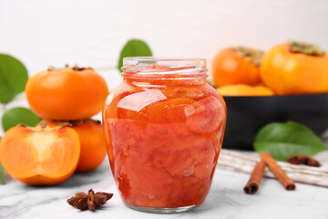 Jar of tasty persimmon jam and ingredients on white marble table