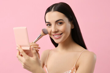 Beautiful woman with cosmetic pocket mirror applying makeup on pink background