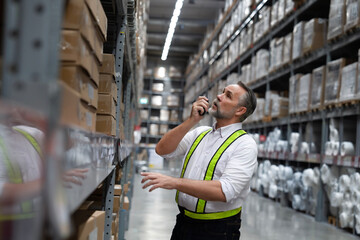 Senior warehouse manager using a walkie-talkie to talk with his team, looking for damaged boxes on shelves. Check shelf number to remove for inspection. Warehouse, inventory, logistic, storage concept