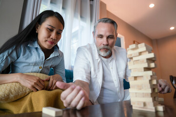 Hispanic husband rolls the dice, playing wood board game with Asian wife in a living room, remove piece of block from tower. Enjoy family activities of recreation at home during free time.