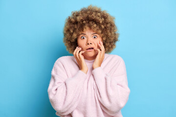 Fototapeta na wymiar Emotional adult woman with curly hair stares in shocked and nervous grabs face, has eyes popped out, dressed in pink pullover, poses over blue background. Human reaction concept