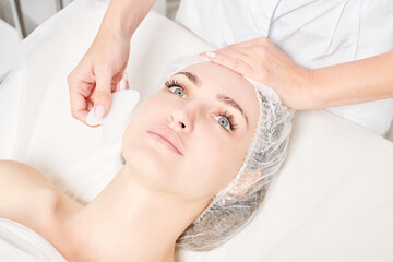 Cosmetologist making facial massage with Gua Sha stone of woman face skin for lymphatic drainage