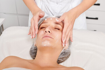 Cosmetologist rubbing moisturizing cream mask into woman face skin for rejuvenation in beauty salon