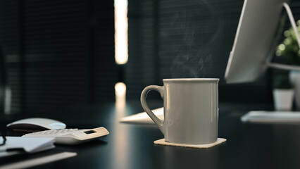 Cup of hot coffee on black wooden table with computer, document and calculator on background. Relaxing moment concept