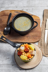 Cooked (Cocido, Guiso) stew, chickpeas with chorizo, bacon, carrot and potato in green saucepan, black casserole with broth and on wooden boards.Concept of traditional Spanish cuisine.