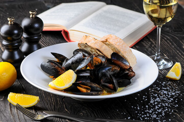 boiled mussels in a plate with lemon and bruschetta on a table with a book and a glass of wine
