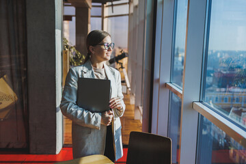 Cheerful young female freelancer in casual clothes standing at a wooden table with a netbook and writing notes while working on a project. Modern young business lady in the office