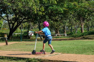 Portrait of active little girl riding scooter on street in outdoor park on summer day. Happy Asian girl wearing a helmet riding a kick scooter in the park. Active leisure and outdoor sport for child.